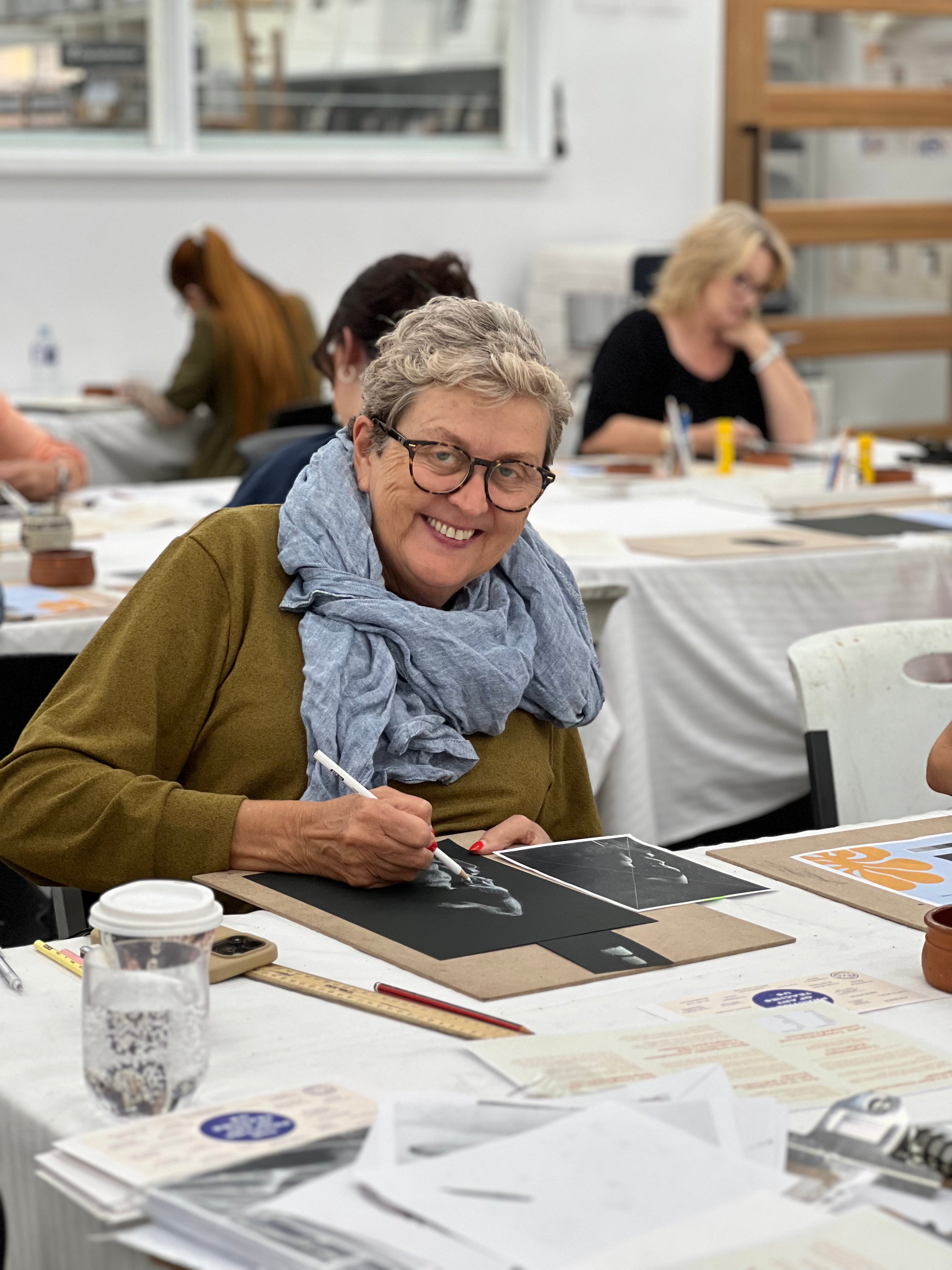 Woman sitting at a table in a workshop or classroom setting, smiling and writing.
